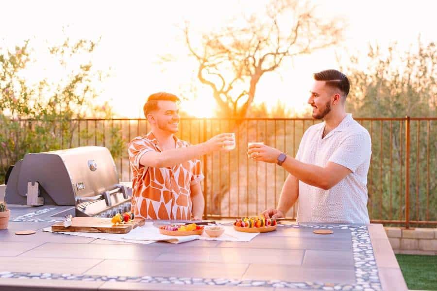 Two men saying cheers at an outdoor kitchen with vegetable kabobs on countertop