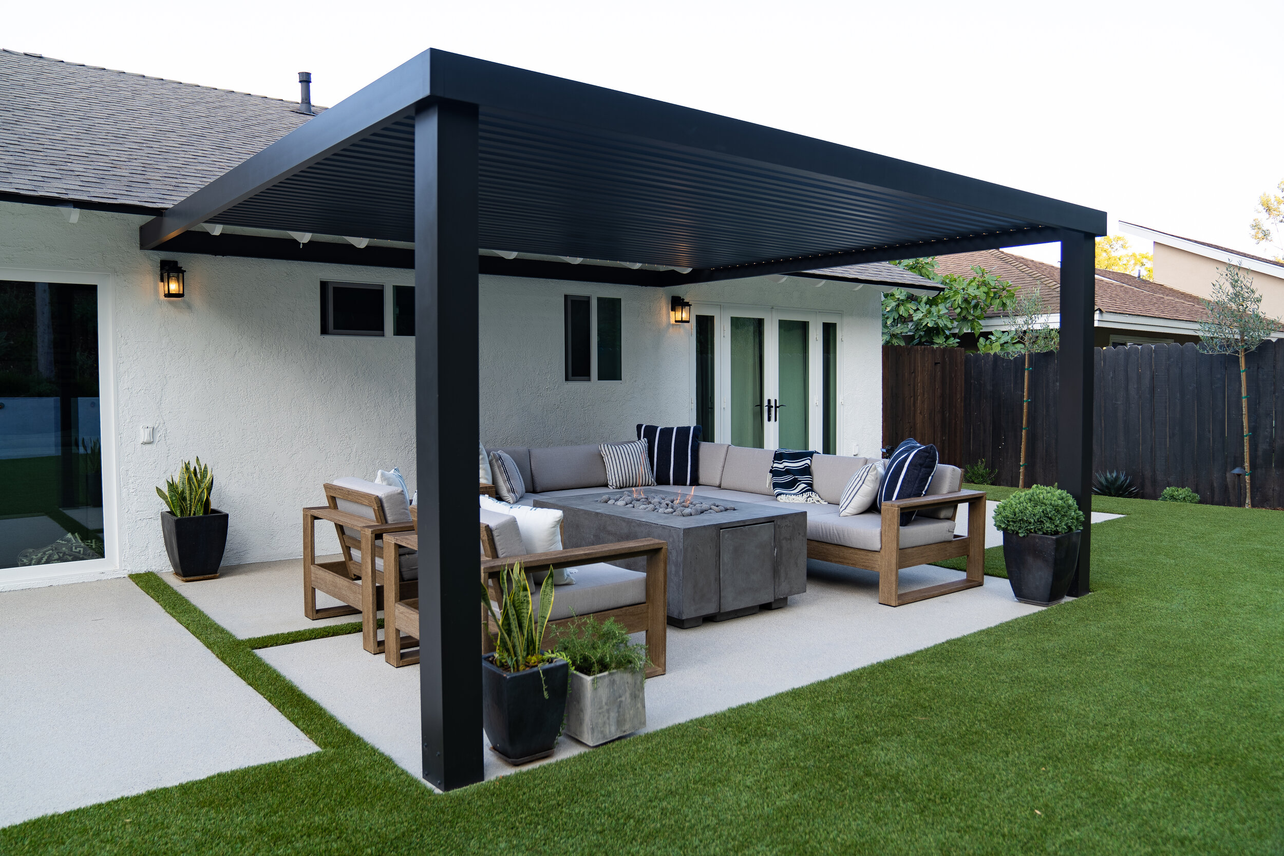 A backyard social area with fire pit and lounge couches covered with a pergola next to a lawn