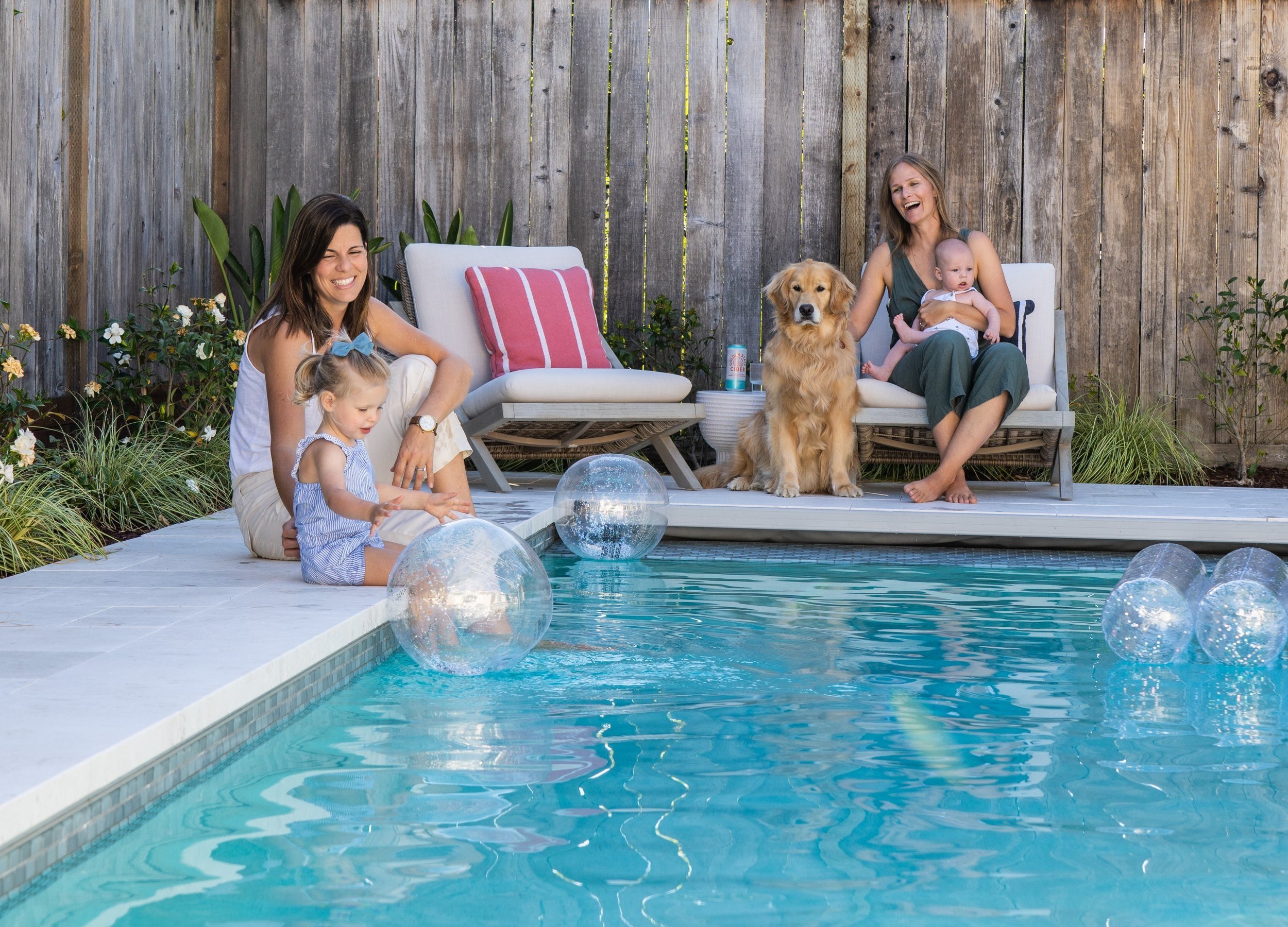 Two women, a girl, baby and dog sitting by a pool in a fenced space