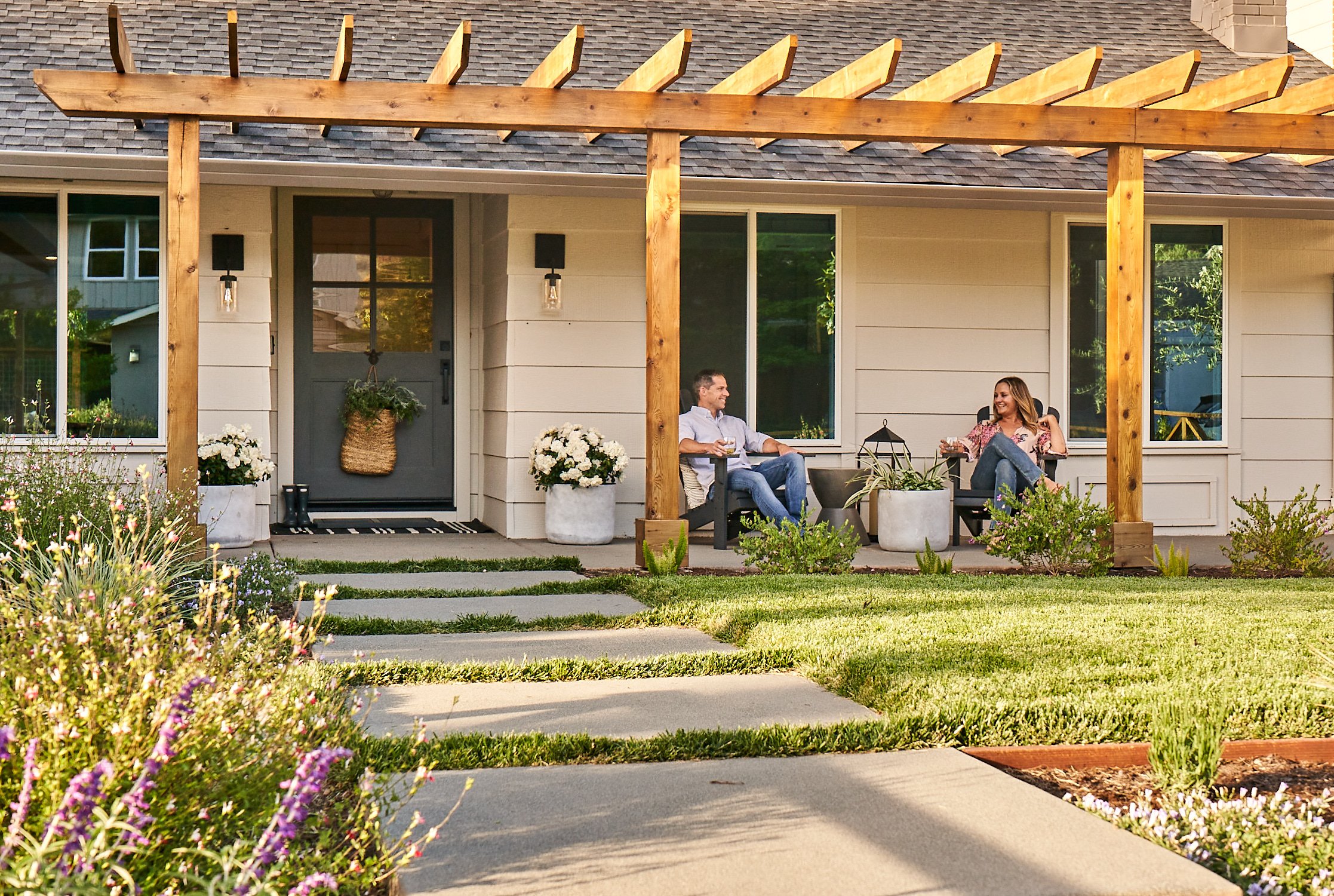 Man and woman sitting on front porch with wooden overhead pergola attached to home