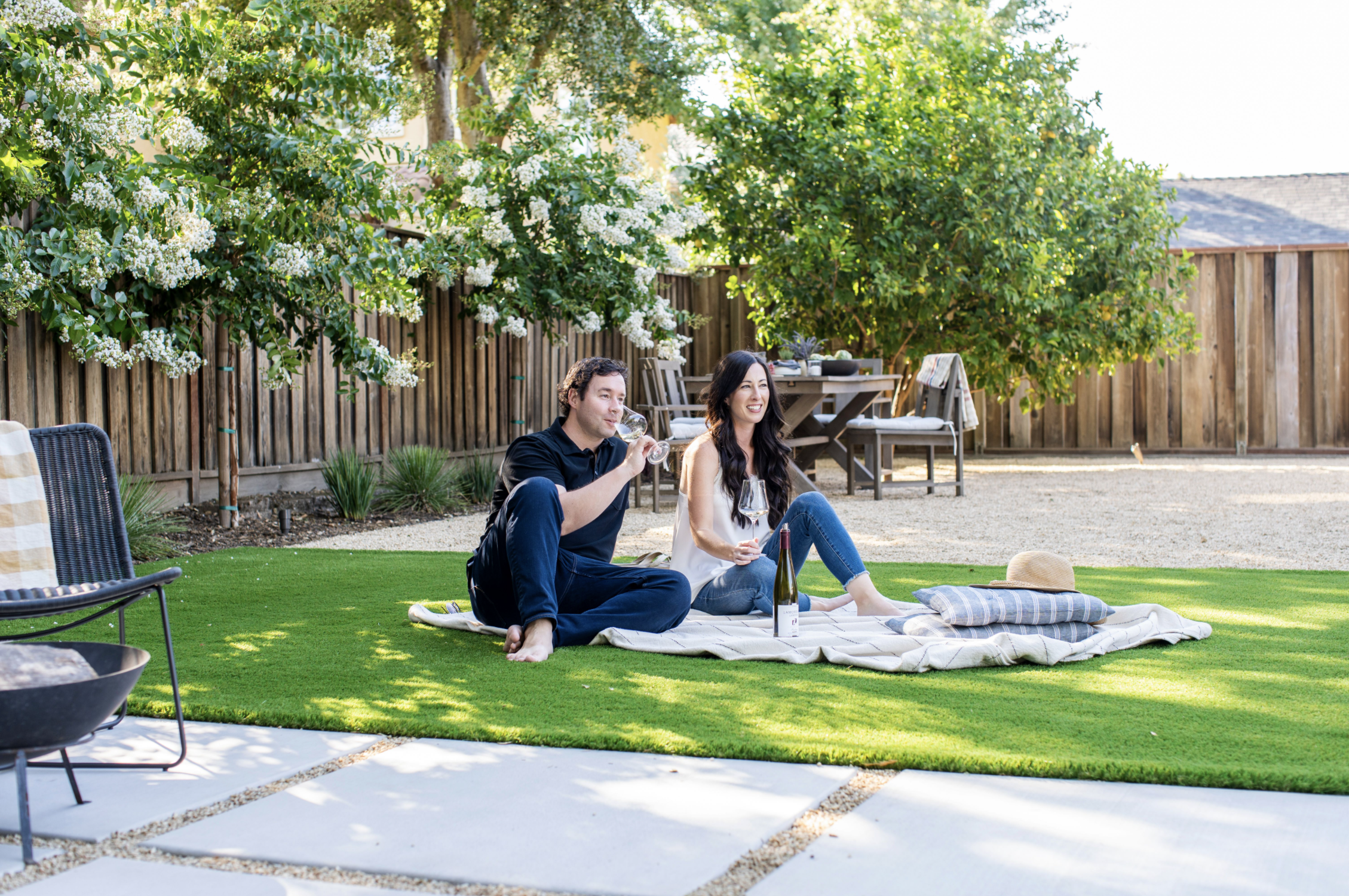 A man and woman having picnic on a lawn in a fenced backyard with, paver stones and dining area