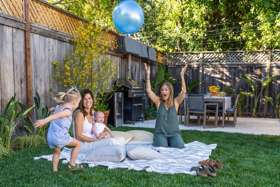 Two women with toddler and baby on a blanket on their backyard lawn with grill and refrigerator on the patio behind them