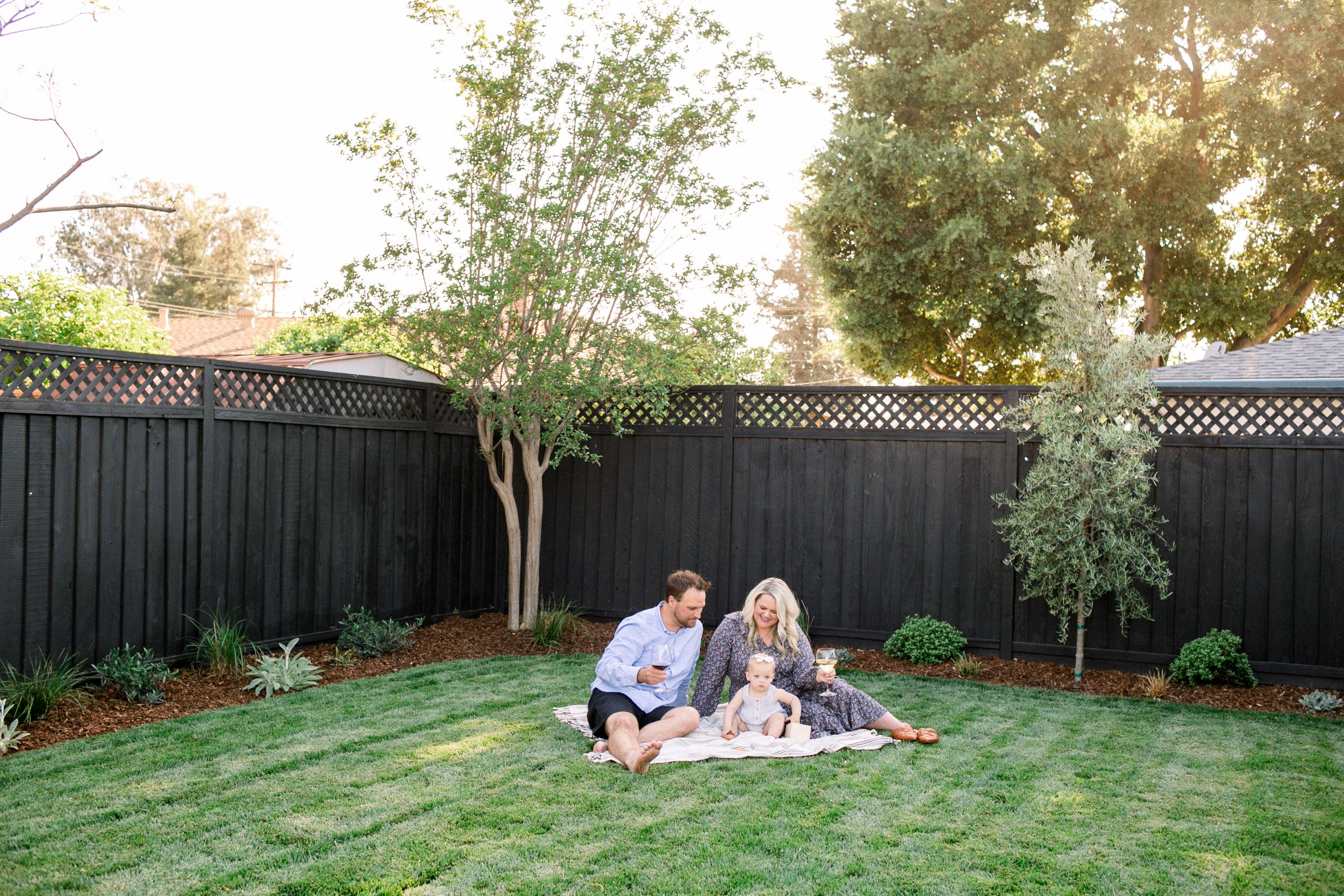 A family of 3 sitting on a lawn in a fenced yard with plants