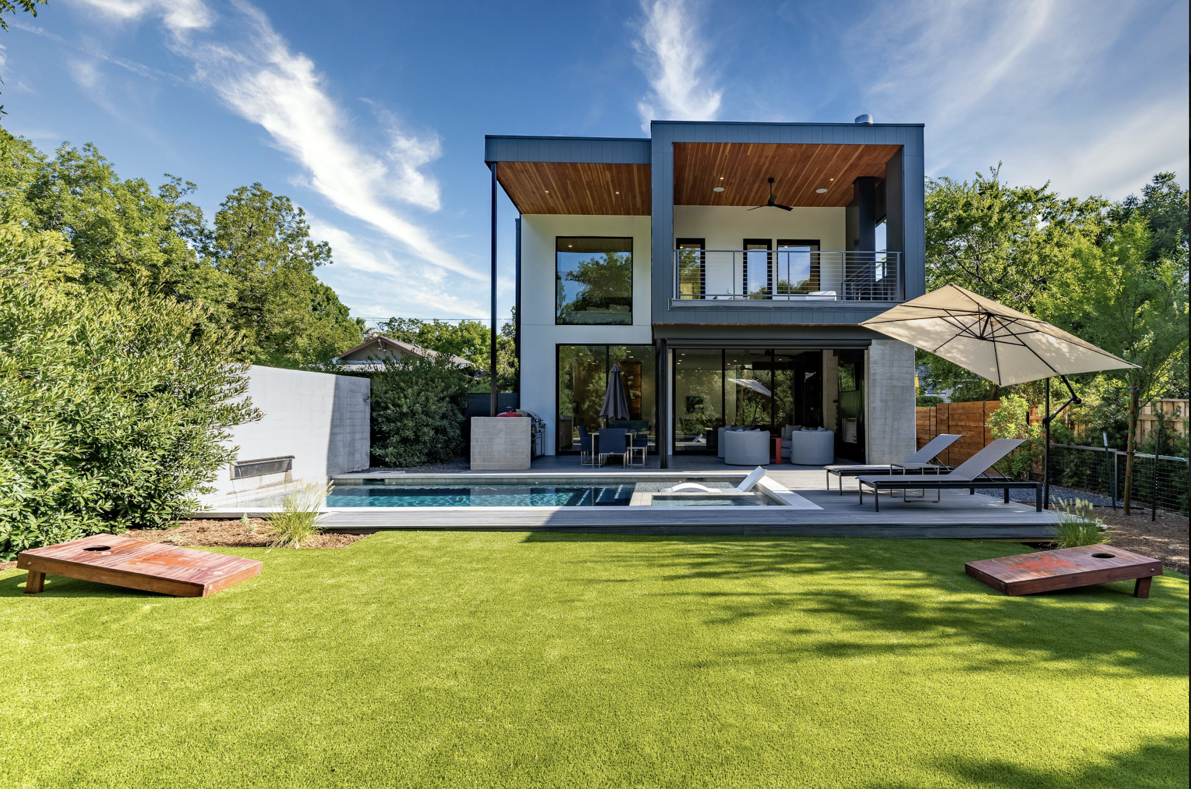 view from the rear of a back yard toward a modern home with second-floor balcony overlooking a pool with a deck lounge chairs, and cantilever umbrella for shade