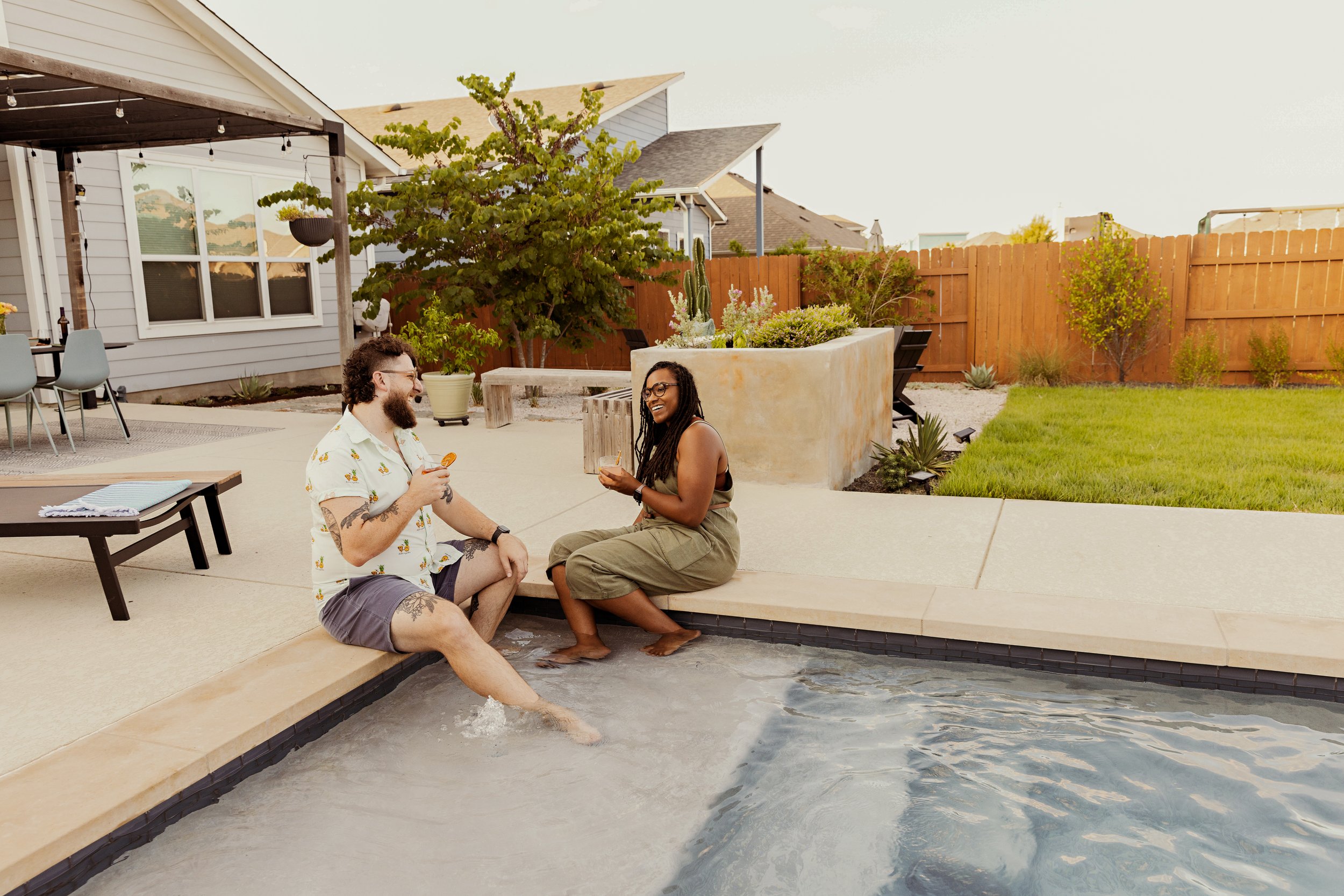 Fenced house, couple relaxing on pool side, tall grass, plants, outdoor chairs, lightings.