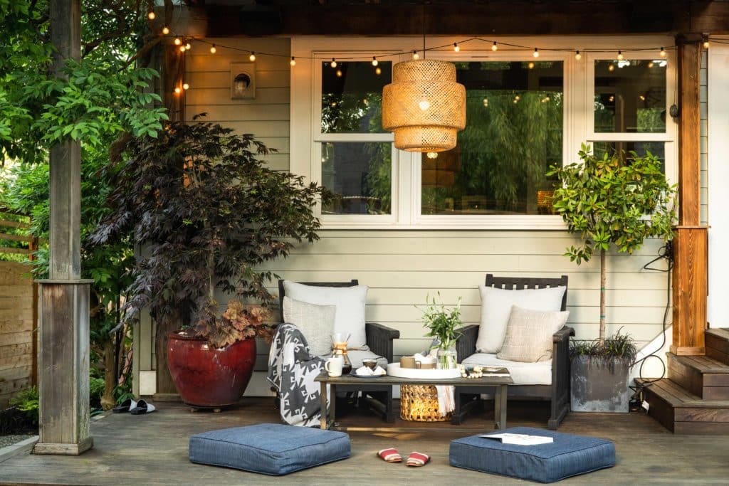 Close up of finished back deck with comfy lounge chairs, coffee table, floor cushions and natural style hanging light and string lights above.