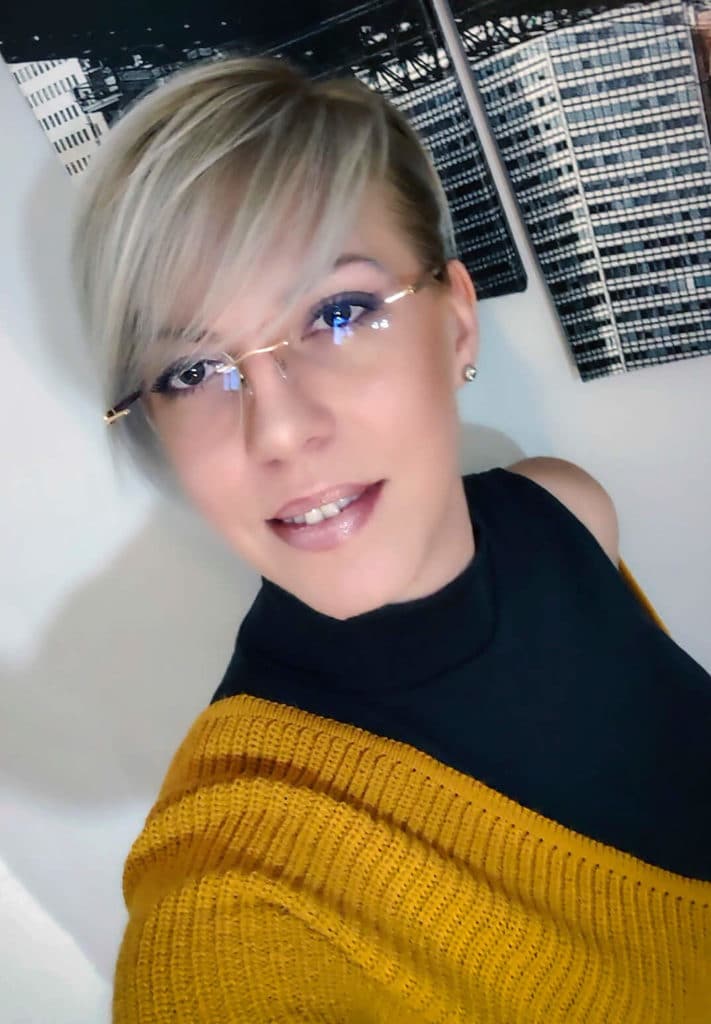Lady wearing glasses, a black sleeveless turtle neck and a mustard cardigan