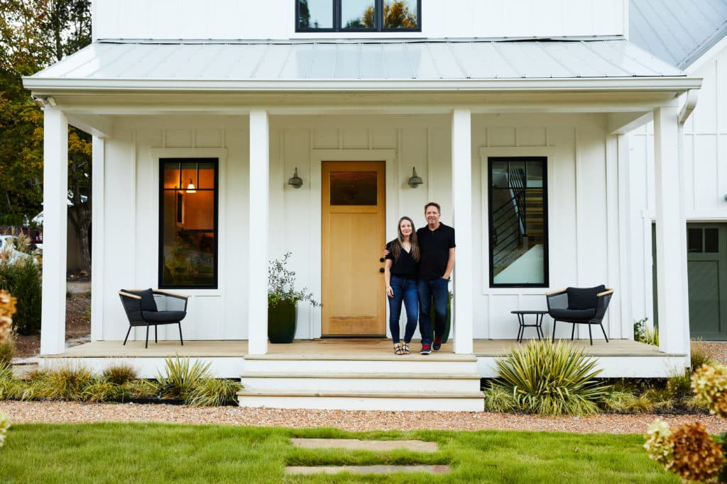 Couple standing on front porch of modern farmhouse style home with planting beds and gravel surrounding porch
