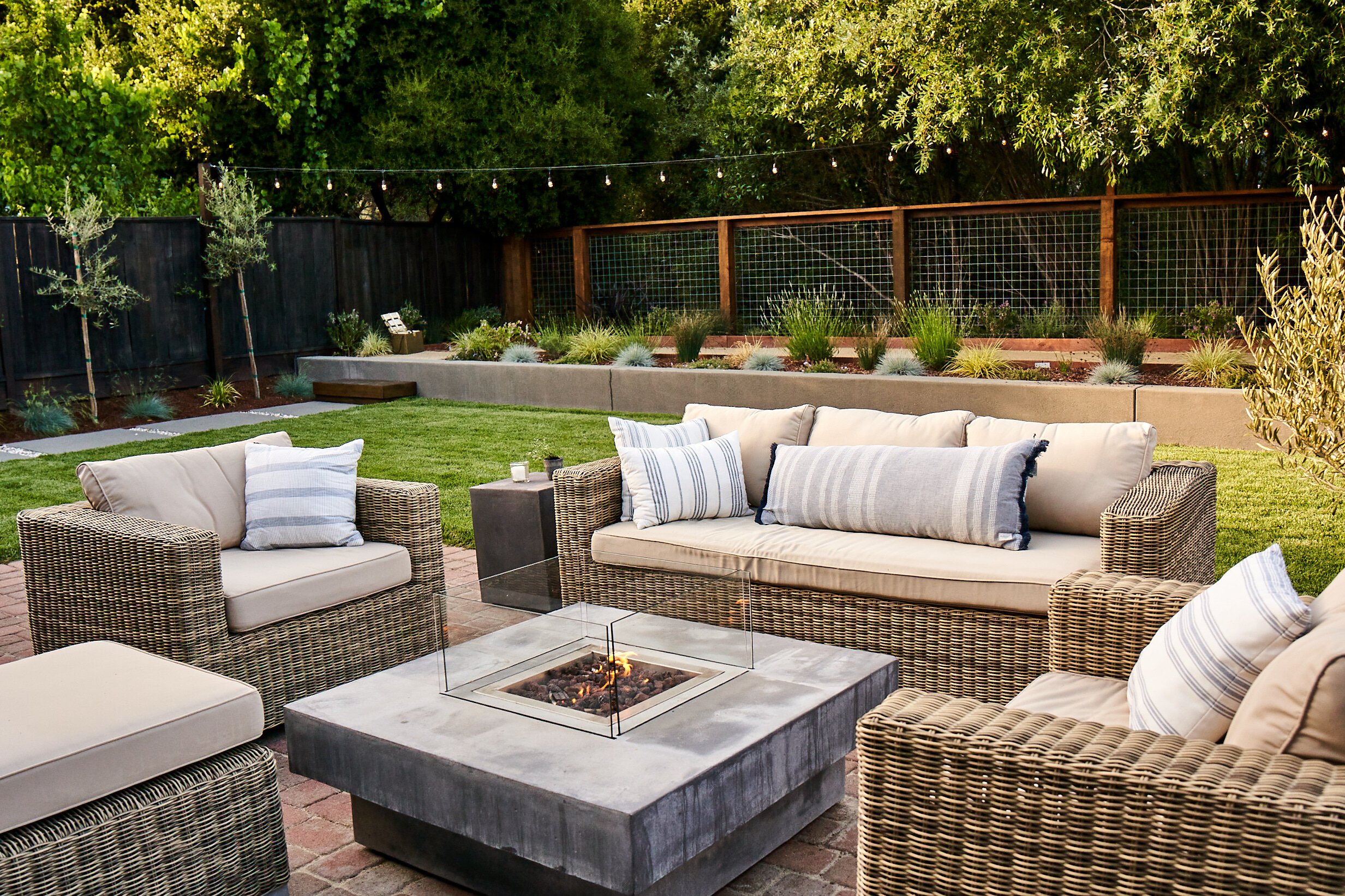 Wicker outdoor seating set on a brick patio surrounding a modern square fire pit with lawn and planting beds behind