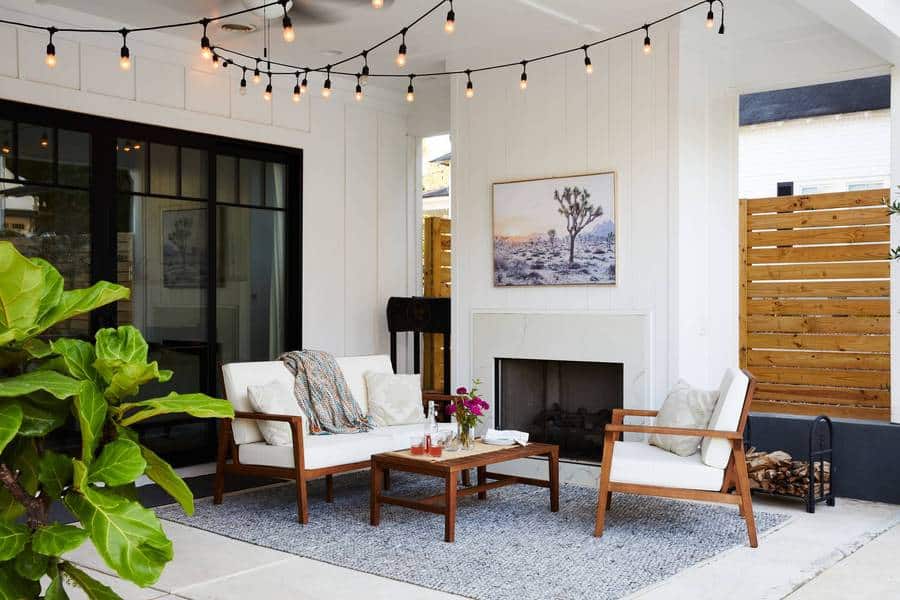 Covered outdoor living room and fireplace with string lights above, otudoor sofa, coffee table, and lounge chair with white cushions on a blue outdoor rug, and fiddle leaf fig in foreground