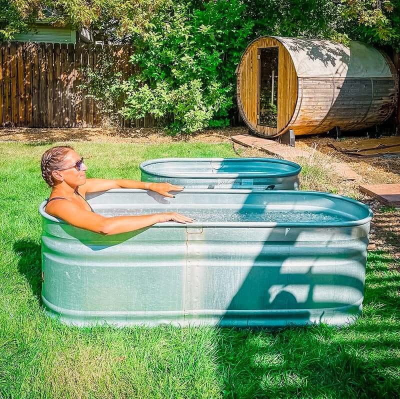 Woman in a stock tank pool in backyard with sauna in background.