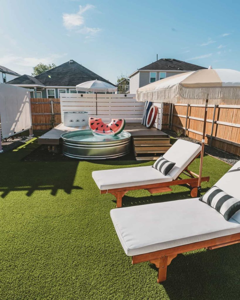 Backyard with artificial turf, lounge chairs and umbrella, and stock tank pool with wooden pool deck.