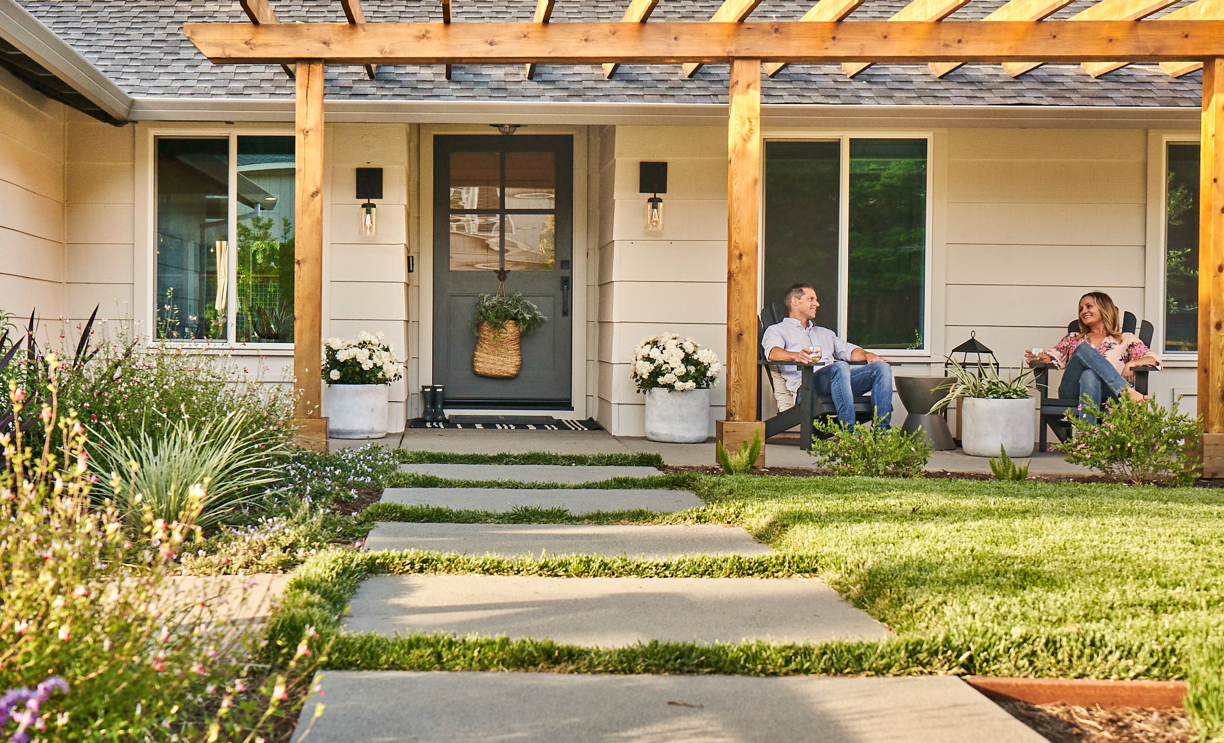 Man and woman sitting on front porch with overhead wooden pergola and paver walkway across front lawn