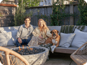 Couple sits with their dog on a concrete bench with a fire pit in front.