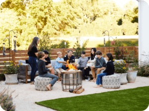Family sits around a fire pit with wood fence behind and plants around.