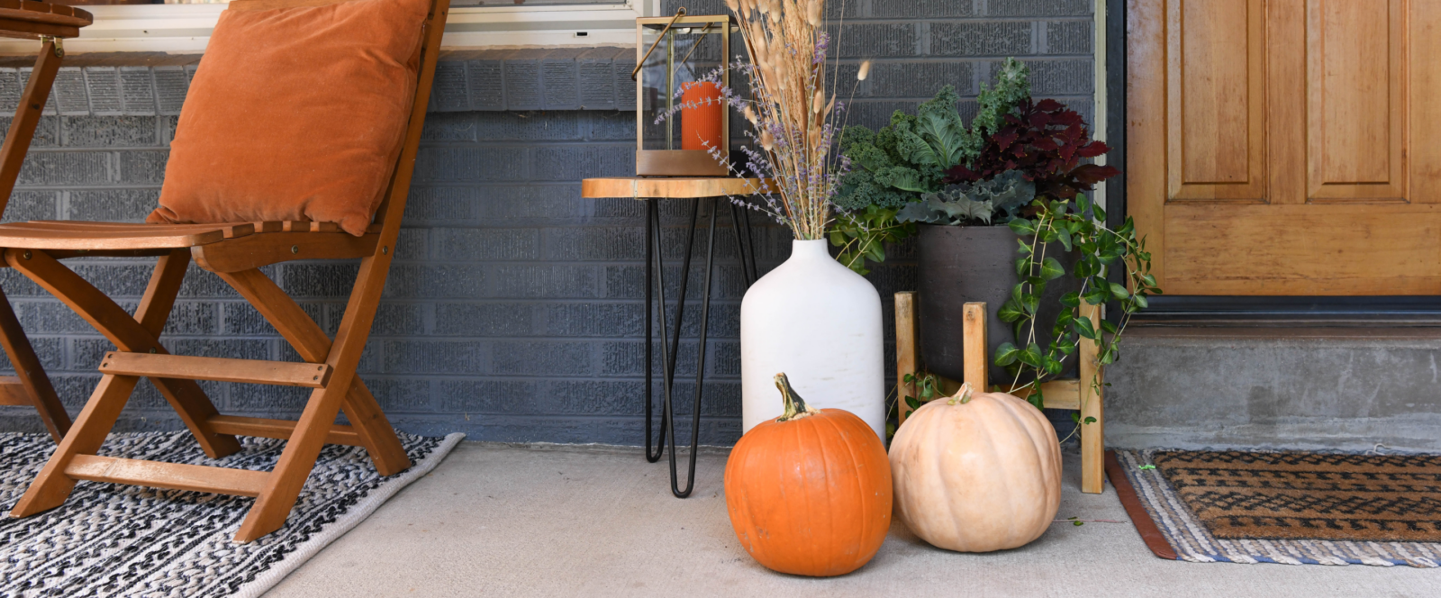 Image of a front porch decorated for fall with pumpkings and a vase with dried plants.