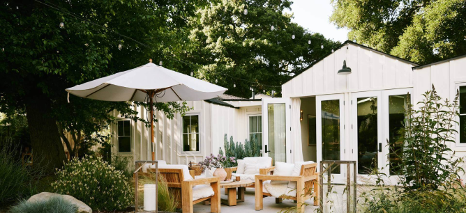 Yard with lots of green and wood outdoor furniture with an outdoor umbrella and white house.