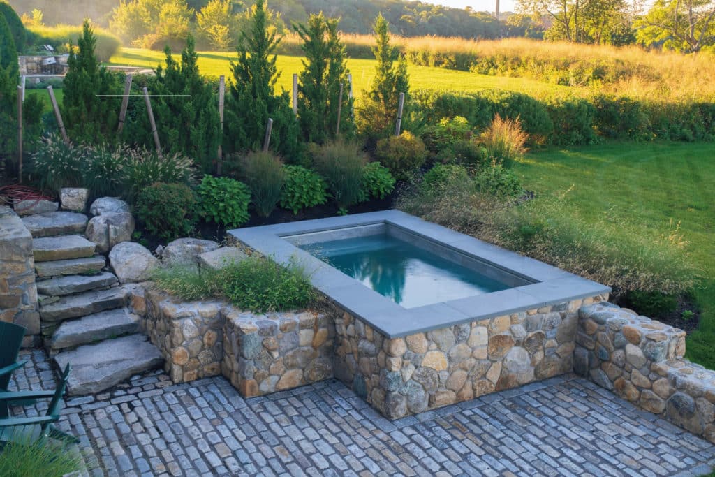 Above ground plunge pool with rustic stone finish and pastoral garden and lawn in background.