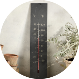 Slate grey outdoor thermometer leaning against a wall with plants.