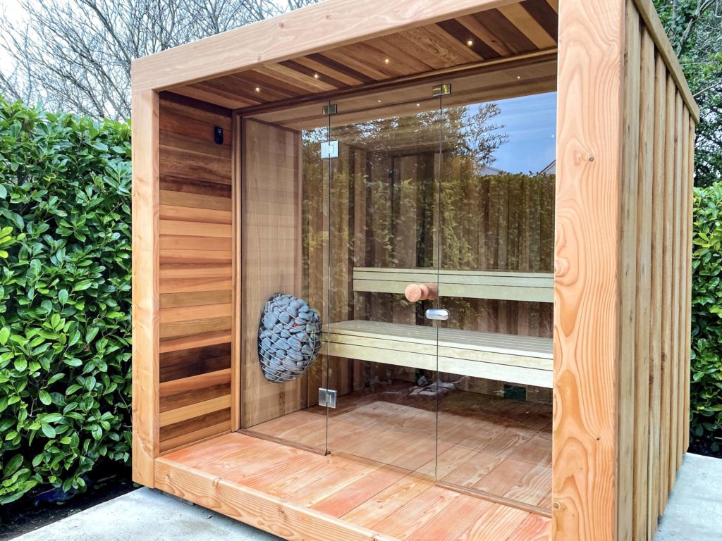 Large, stylish outdoor sauna with tiered benches and cedar wood construction with teardrop-shaped heater filled with sauna stones fixed to interior wall.