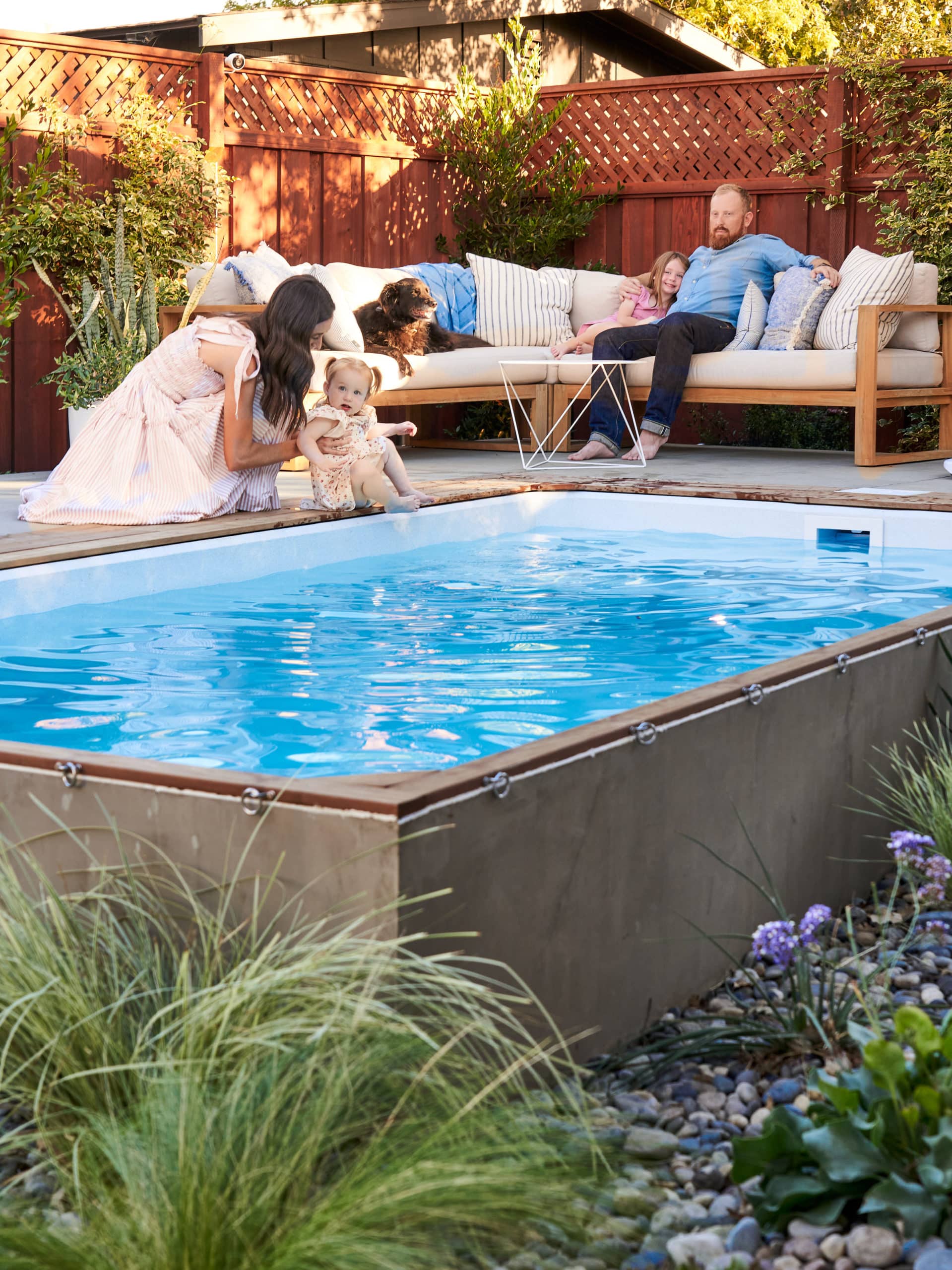 Man and woman with two young children and dog on a concrete patio with sectional around a plunge pool.