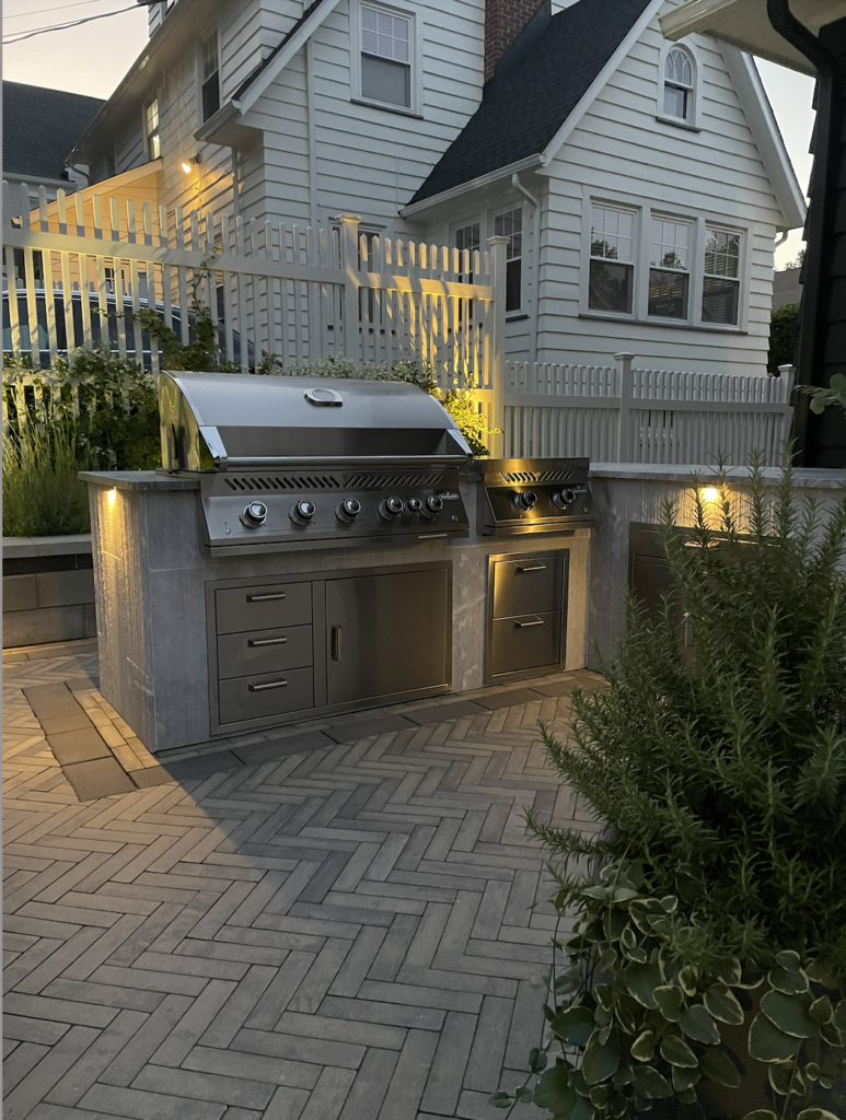 An outdoor kitchen with a grill, lit for dusk
