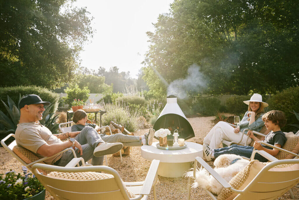 Family sits around an outdoor fireplace in white chairs. They are surrounded by gravel hardscaping and lush California native plants.
