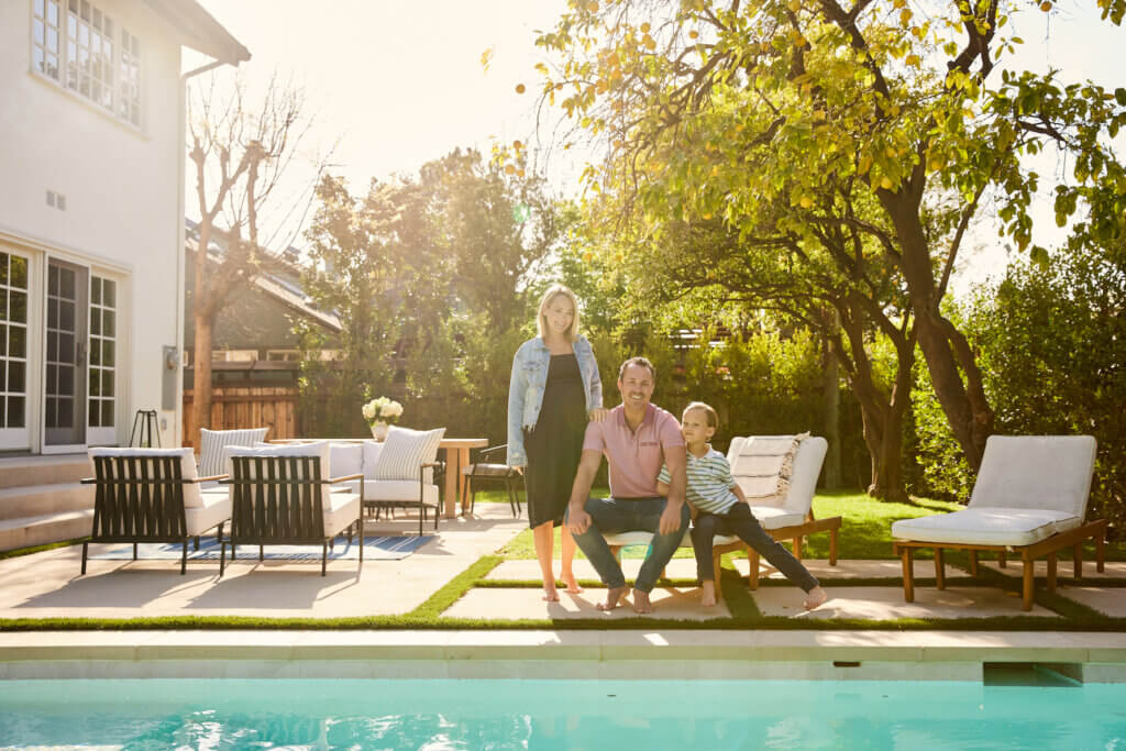Woman, man and child sit together on the concrete deck of their pool. They are flanked by an outdoor living room, chaise lounges and a large lemon tree.