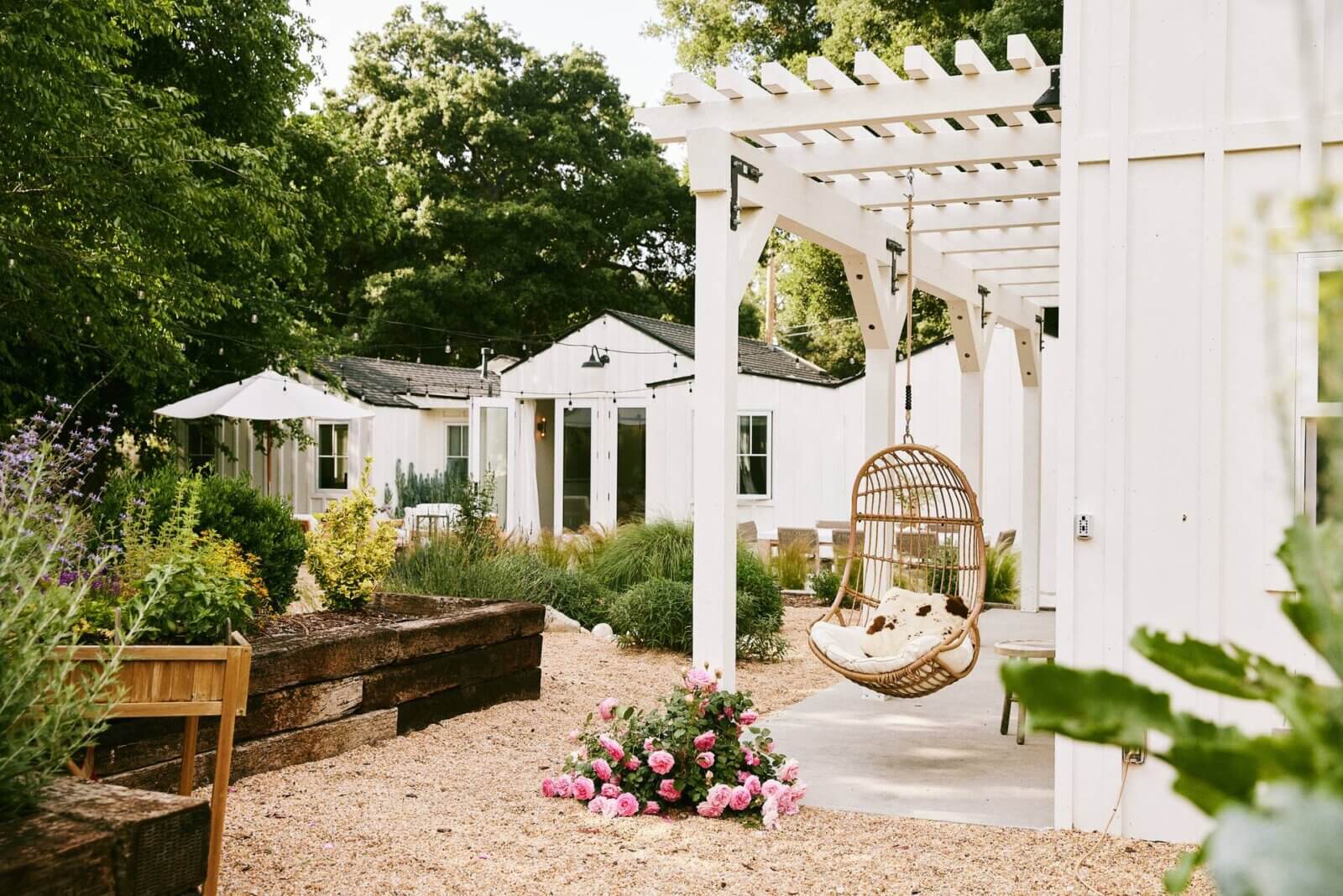 Modern farmhouse with white pergola and hanging chair in lush backyard by Yardzen