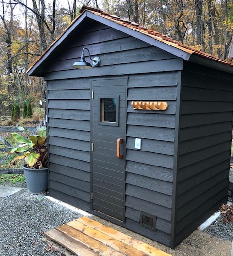 Modern black-painted wooden outdoor sauna with black metal door with small window, and towel hooks and wall light on the facade.